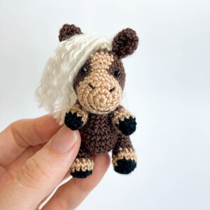 May include: A small, handmade crochet horse figurine. The horse features a brown body, tan face, and black hooves. It has a white, fluffy mane and dark brown ears. The horse is a miniature, handcrafted item.