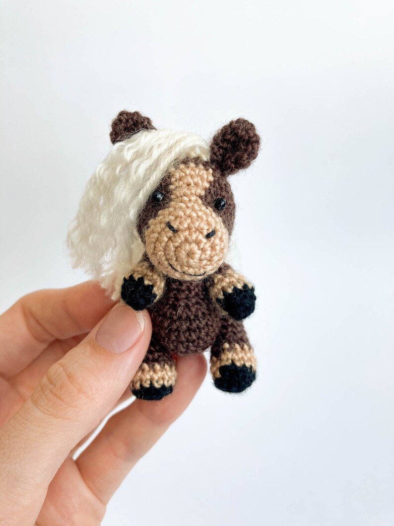 May include: A small, handmade crochet horse figurine. The horse features a brown body, tan face, and black hooves. It has a white, fluffy mane and dark brown ears. The horse is a miniature, handcrafted item.