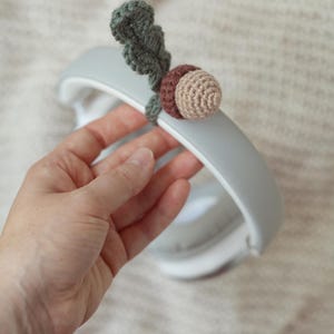 May include: A gray headband with a crocheted acorn charm attached. The acorn is brown and beige with a green leaf.