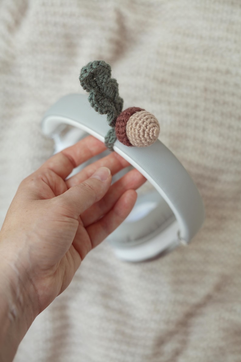 May include: A gray headband with a crocheted acorn charm attached. The acorn is brown and beige with a green leaf.