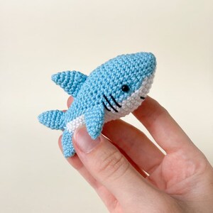 May include: A small crocheted blue shark with white detailing and black eyes. The shark is being held in a hand.