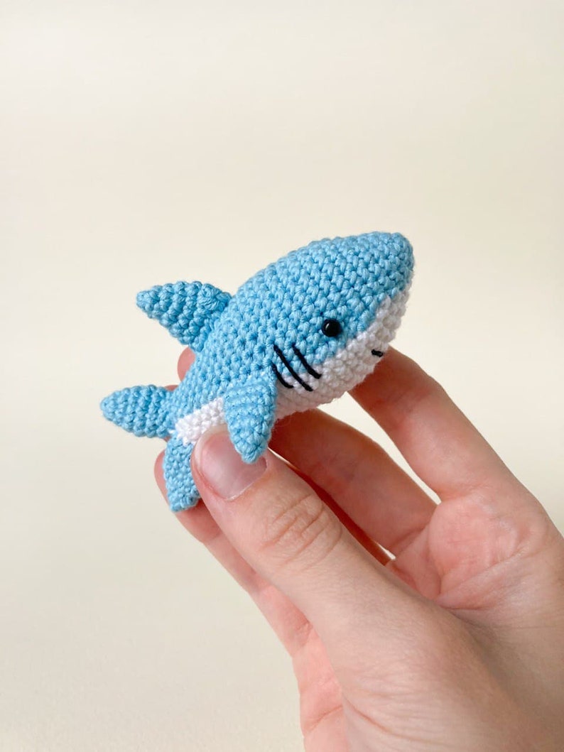 May include: A small crocheted blue shark with white detailing and black eyes. The shark is being held in a hand.