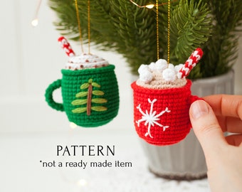 May include: Two crocheted hot chocolate mug ornaments, one green with a Christmas tree design and one red with a snowflake design.  The ornaments are hanging from a Christmas tree.