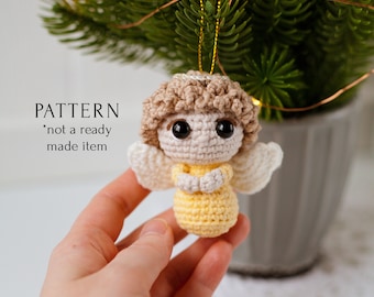 Op de afbeelding: Een gehaakt engel-ornament met een gele jurk, witte vleugels en bruin haar. De engel hangt aan een gouden touwtje en wordt vastgehouden door een hand.