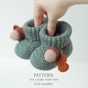 May include: A pair of green crocheted baby booties with acorn accents. The booties are held in a hand. The text "PATTERN *not a ready made item 0-12 months" is below the booties.