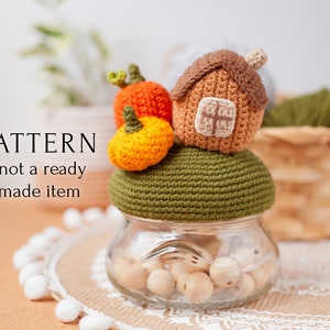 Op de afbeelding: Haakpatroon voor een herfst-thema deksel voor een pot met een klein huis, pompoenen en een groene basis. De pot is gevuld met houten kralen. De tekst "PATTERN *not a ready made item" is zichtbaar.