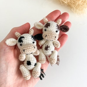 May include: Two small crocheted cows, one white with black spots and one white with brown spots, are held in a person's hand. The cows have black eyes and brown noses.