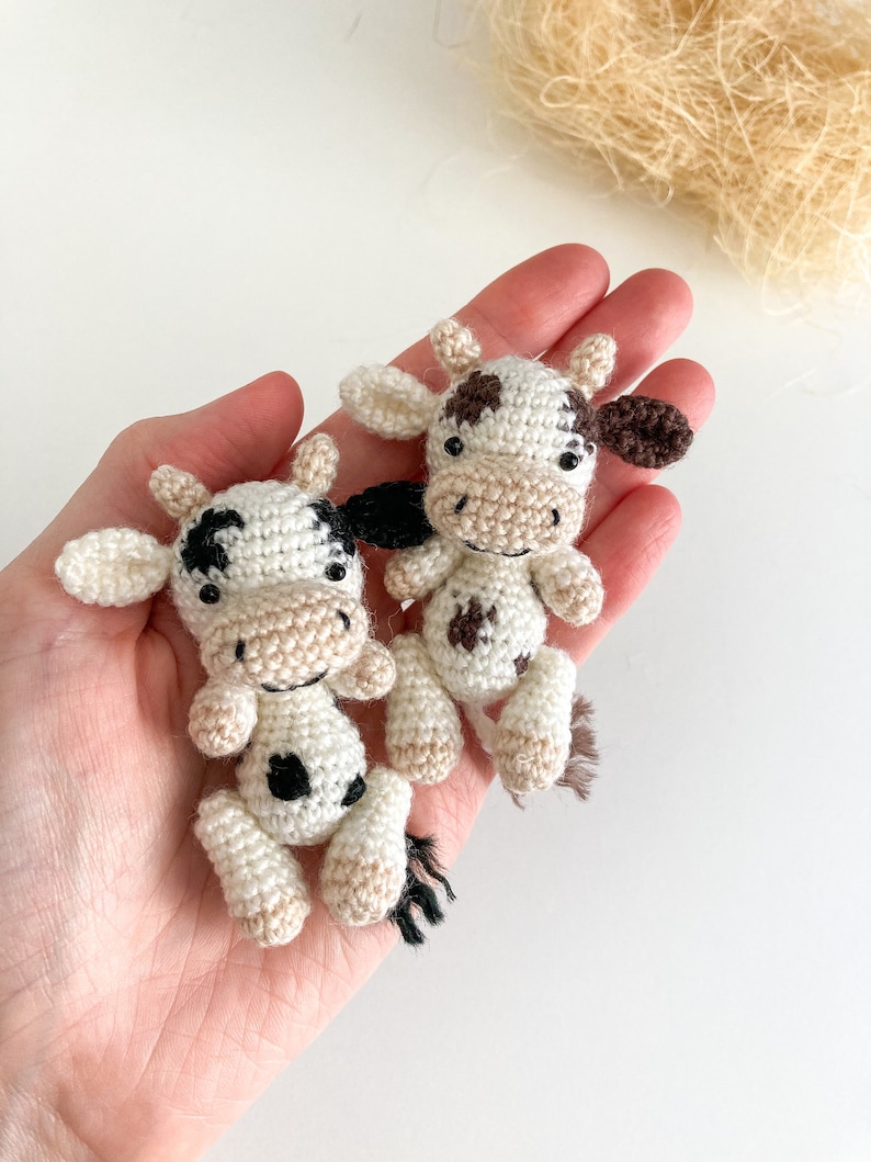 May include: Two small crocheted cows, one white with black spots and one white with brown spots, are held in a person's hand. The cows have black eyes and brown noses.