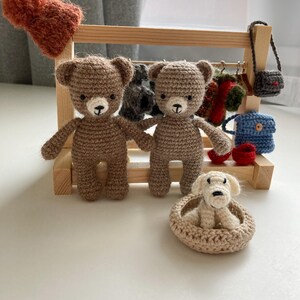 May include: Two brown crocheted teddy bears standing on a wooden shelf with a white crocheted dog in a bed. The shelf has a variety of crocheted accessories hanging from it.