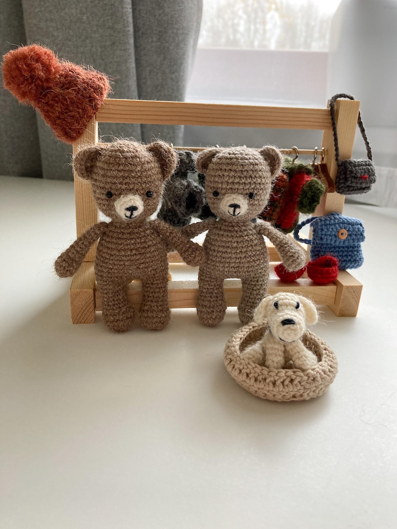 May include: Two brown crocheted teddy bears standing on a wooden shelf with a white crocheted dog in a bed. The shelf has a variety of crocheted accessories hanging from it.