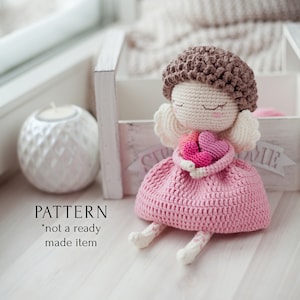 Puede incluir: Una muñeca de ángel de crochet con cabello castaño, vestido rosa y alas blancas que sostiene tres corazones rosas. La muñeca está sentada sobre una superficie de madera blanca. PATTERN *not a ready made item*