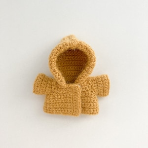 May include: A small, crocheted hooded jacket in a mustard yellow color. The jacket has a simple design with a button closure and short sleeves.
