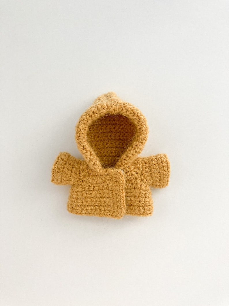 May include: A small, crocheted hooded jacket in a mustard yellow color. The jacket has a simple design with a button closure and short sleeves.