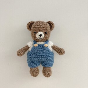 May include: A brown crocheted teddy bear wearing blue overalls with white buttons and a white shirt. The bear has black eyes and a brown nose.