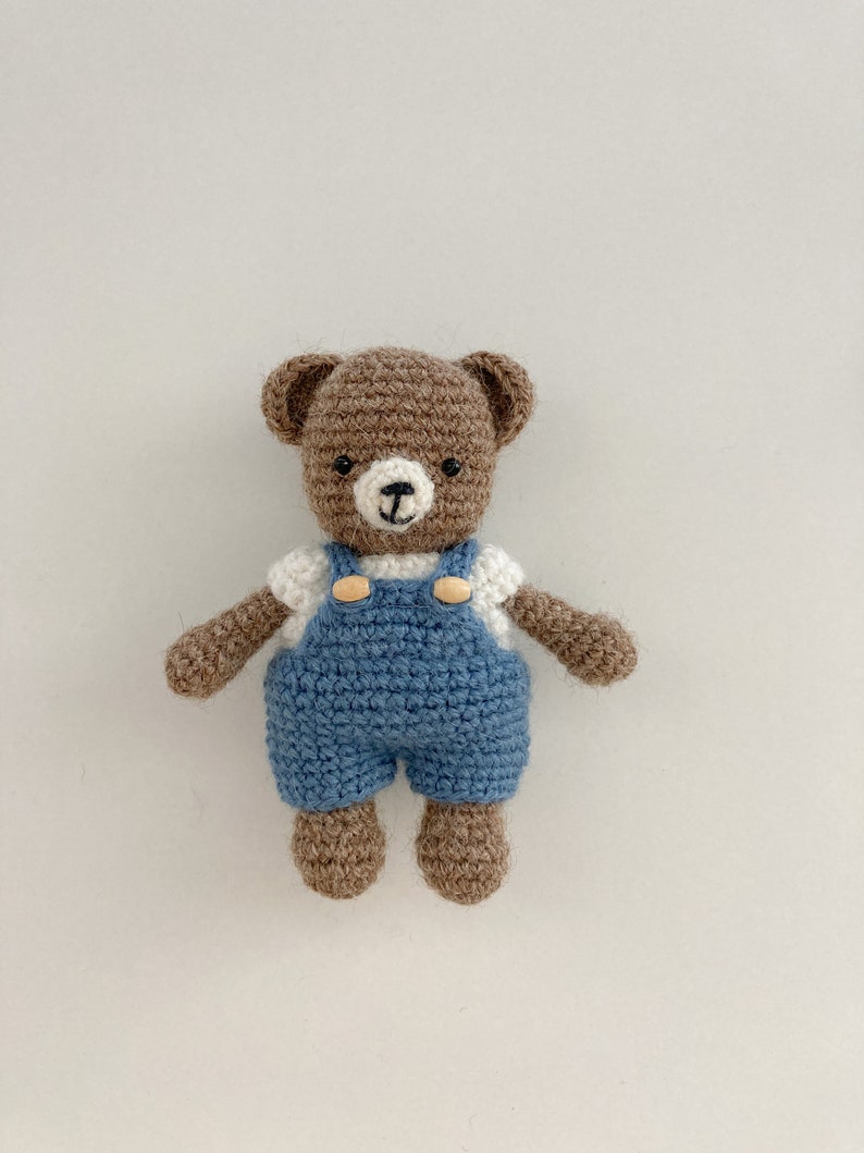 May include: A brown crocheted teddy bear wearing blue overalls with white buttons and a white shirt. The bear has black eyes and a brown nose.