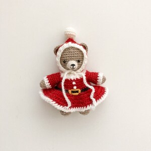 K&ouml;nnte beinhalten: Ein geh&auml;kelter Teddyb&auml;r, der ein rotes Weihnachtsmannkost&uuml;m mit wei&szlig;en Verzierungen tr&auml;gt. Der B&auml;r hat ein braunes Gesicht und schwarze Augen.