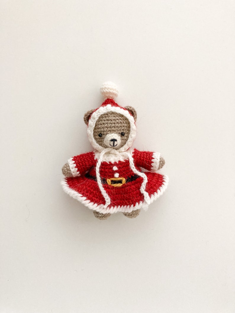K&ouml;nnte beinhalten: Ein geh&auml;kelter Teddyb&auml;r, der ein rotes Weihnachtsmannkost&uuml;m mit wei&szlig;en Verzierungen tr&auml;gt. Der B&auml;r hat ein braunes Gesicht und schwarze Augen.