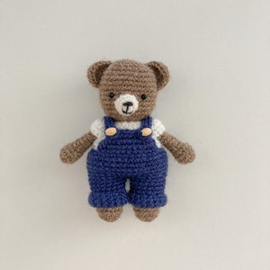 May include: A brown crocheted teddy bear wearing blue overalls with white buttons. The bear has a white face with black eyes and a brown nose.