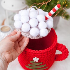 Pu&ograve; includere: Una tazza rossa all'uncinetto con una stella bianca e un design a forma di albero di Natale verde. La tazza ha un coperchio rimovibile con pompon bianchi e una cannuccia a uncinetto rosso e bianca.