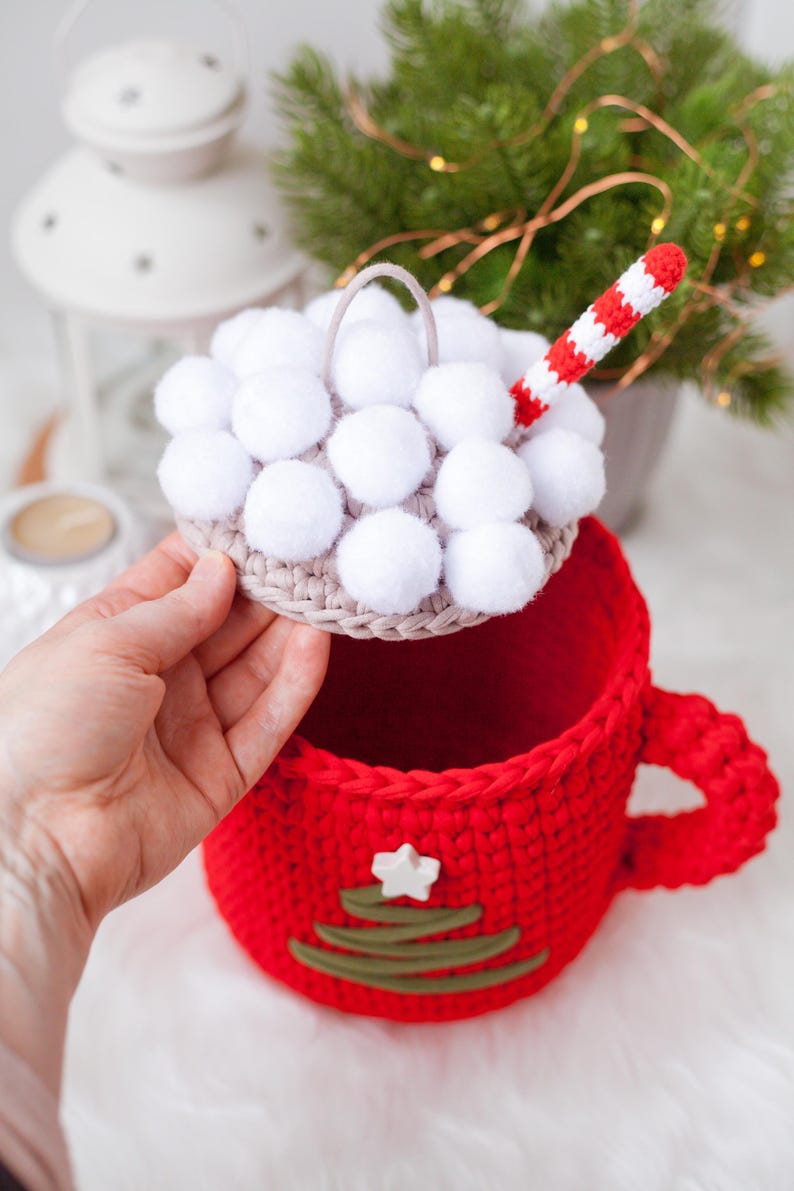 Pu&ograve; includere: Una tazza rossa all'uncinetto con una stella bianca e un design a forma di albero di Natale verde. La tazza ha un coperchio rimovibile con pompon bianchi e una cannuccia a uncinetto rosso e bianca.