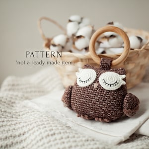 May include: Crochet owl rattle toy with a wooden ring. The owl is brown with white eyes and a closed-eye expression. The toy is a pattern, not a finished product.