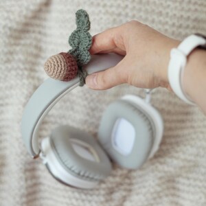 May include: Gray headphones with a crocheted acorn charm attached to the headband. The acorn is brown and green.