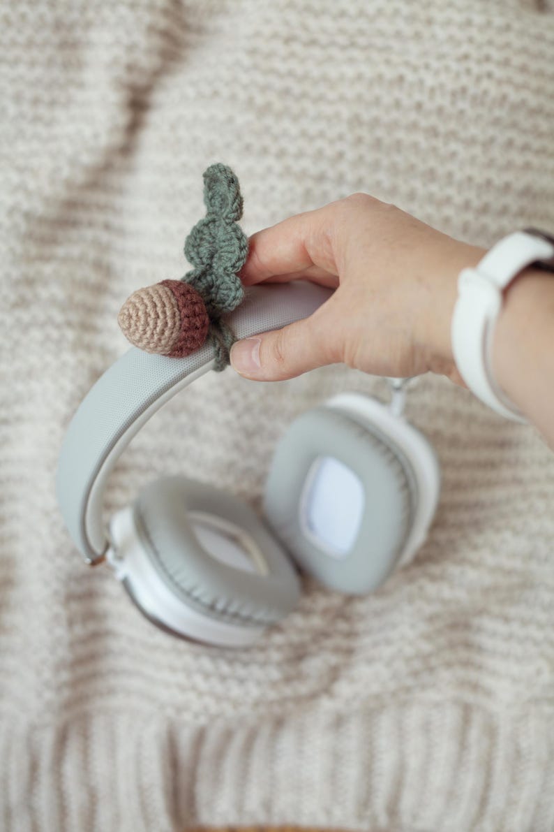 May include: Gray headphones with a crocheted acorn charm attached to the headband. The acorn is brown and green.