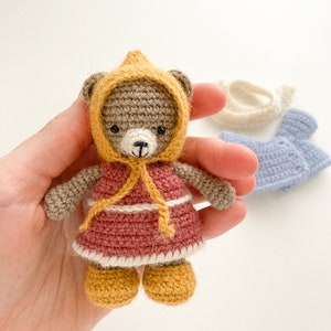 Op de afbeelding: Een gehaakte teddybeer die een gele jurk met capuchon en bruine schoenen draagt. De beer houdt een geel lint in zijn pootjes.