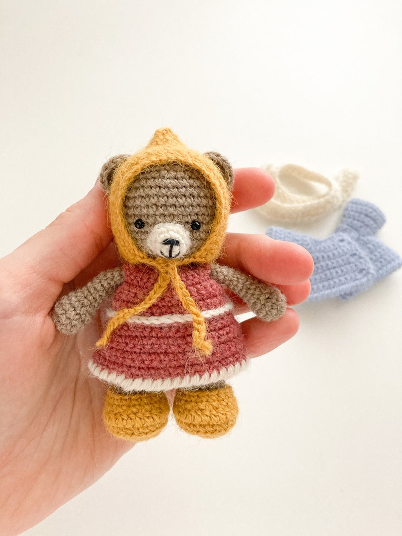 Op de afbeelding: Een gehaakte teddybeer die een gele jurk met capuchon en bruine schoenen draagt. De beer houdt een geel lint in zijn pootjes.