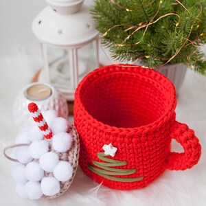 Pu&ograve; includere: Una tazza rossa all'uncinetto con un disegno di albero di Natale verde e una stella bianca. La tazza &egrave; appoggiata su una superficie bianca e soffice accanto a un ornamento bianco all'uncinetto con pompon bianchi.