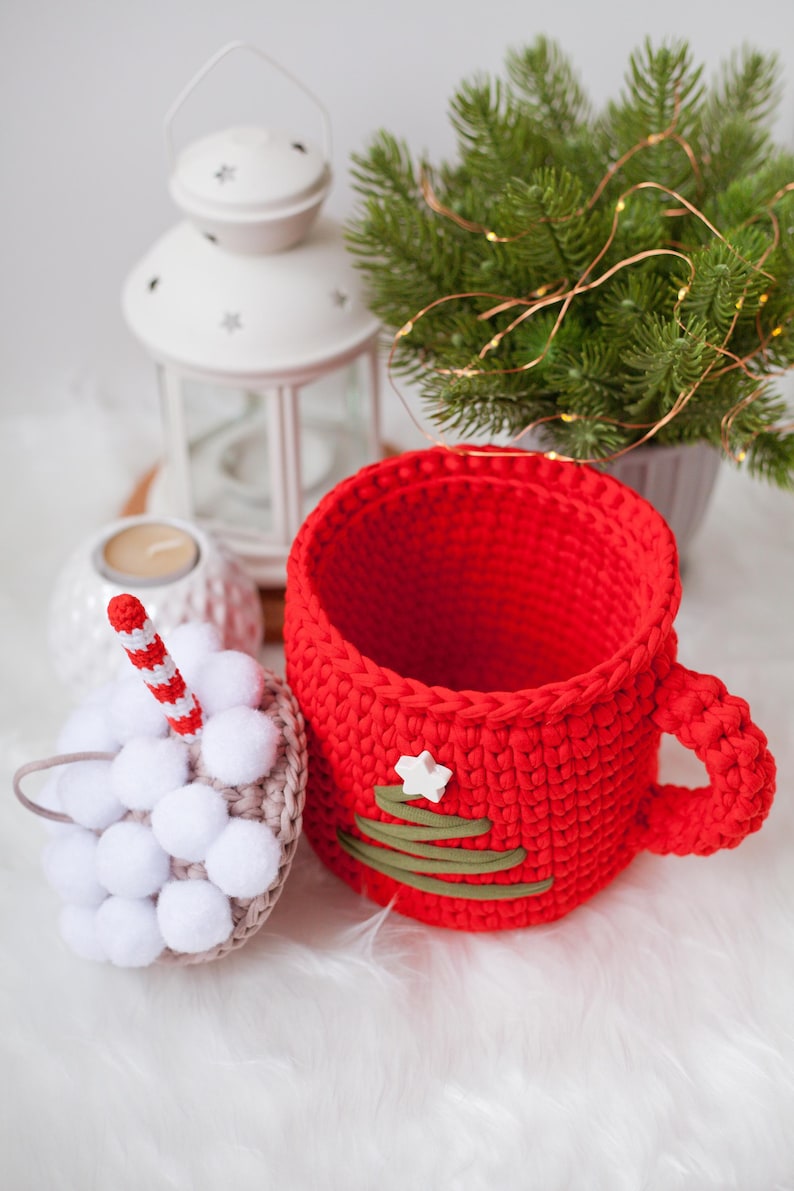 Pu&ograve; includere: Una tazza rossa all'uncinetto con un disegno di albero di Natale verde e una stella bianca. La tazza &egrave; appoggiata su una superficie bianca e soffice accanto a un ornamento bianco all'uncinetto con pompon bianchi.
