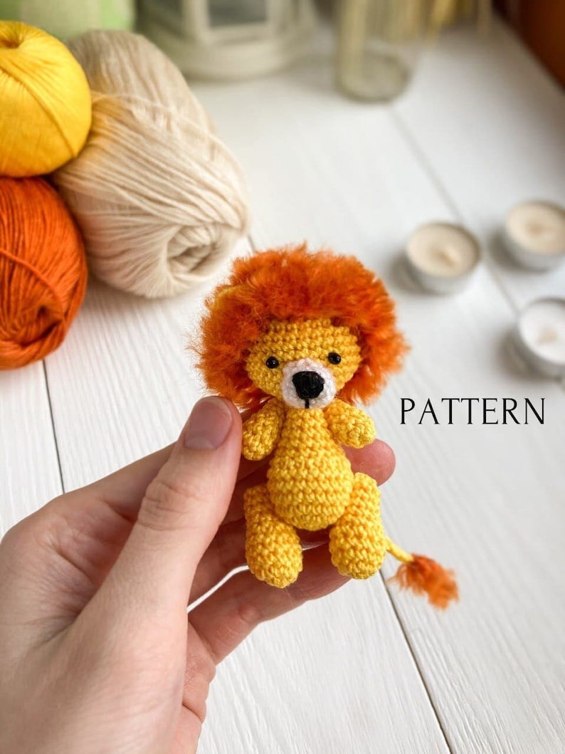 K&ouml;nnte beinhalten: Ein geh&auml;keltes Amigurumi-L&ouml;wenspielzeug mit gelbem K&ouml;rper, orangefarbener M&auml;hne und schwarzen Augen. Das Spielzeug wird in einer Hand gehalten. Das Wort "PATTERN" ist in wei&szlig;er Schrift auf wei&szlig;em Hintergrund geschrieben.