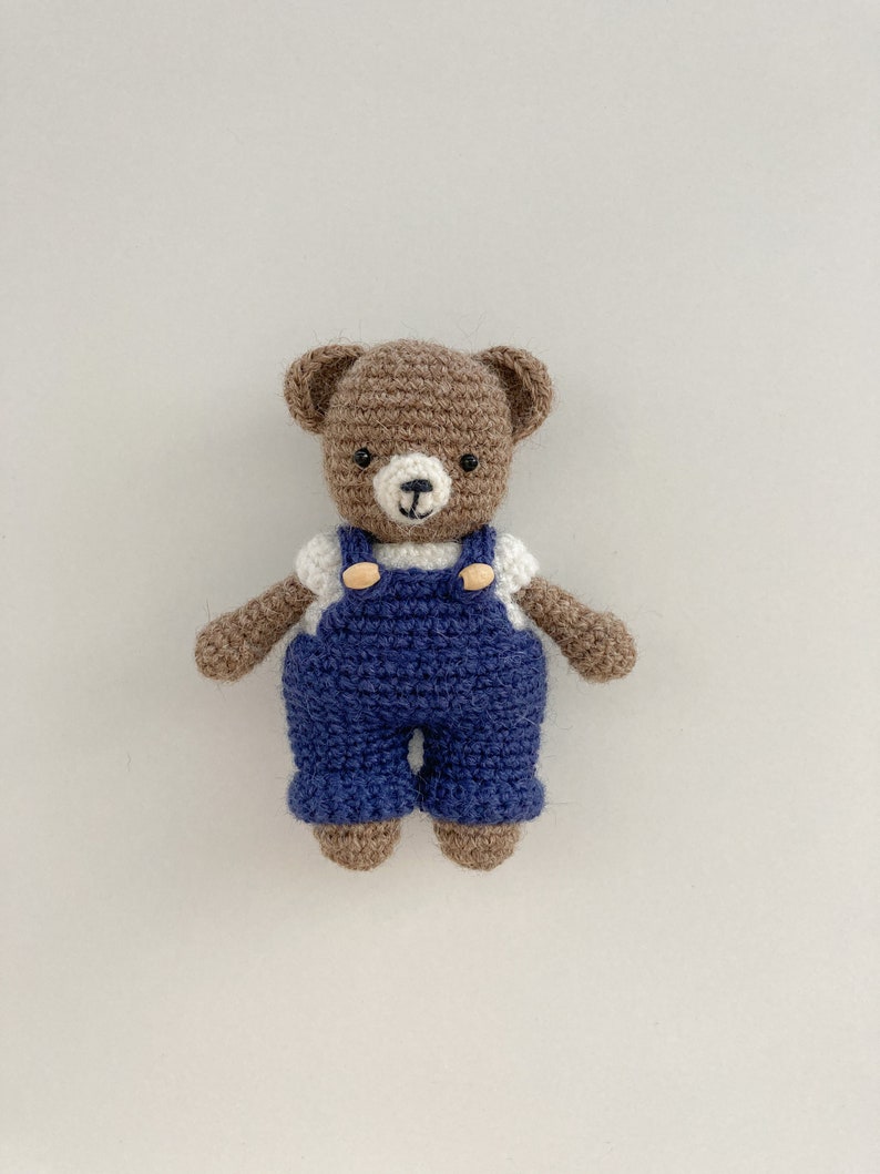 May include: A brown crocheted teddy bear wearing blue dungarees with white buttons. The bear has a white face with black eyes and a brown nose.