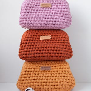 May include: Three crocheted makeup bags stacked on top of each other. The top bag is pink, the middle bag is orange, and the bottom bag is yellow. Each bag has a small brown leather tag with the word "PRIMAVERA" printed on it.