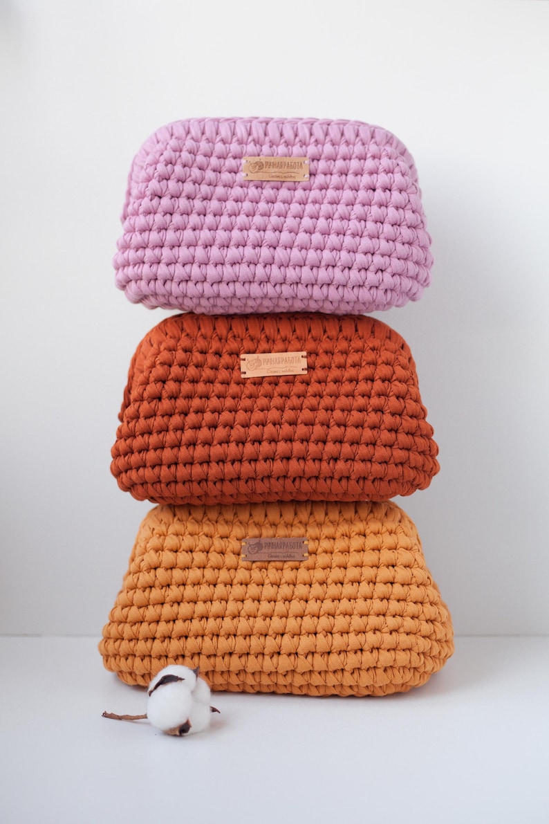 May include: Three crocheted makeup bags stacked on top of each other. The top bag is pink, the middle bag is orange, and the bottom bag is yellow. Each bag has a small brown leather tag with the word "PRIMAVERA" printed on it.
