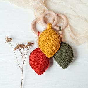 May include: A wooden teether with three crocheted leaf-shaped toys attached. The leaves are yellow, orange, and green.
