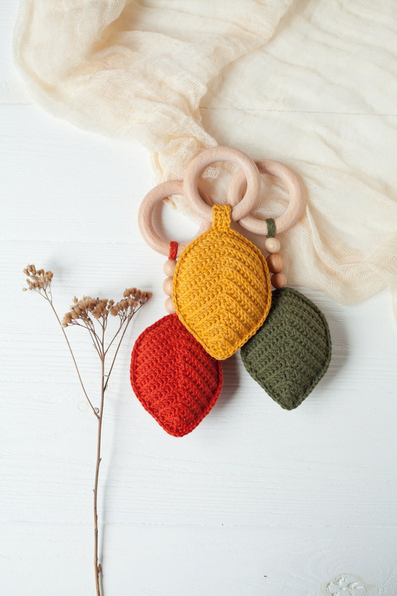 May include: A wooden teether with three crocheted leaf-shaped toys attached. The leaves are yellow, orange, and green.