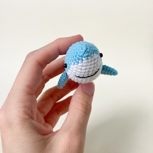 May include: A small crocheted blue whale with white detailing and black eyes. The whale is being held in a hand.