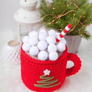 Pu&ograve; includere: Una tazza di uncinetto rossa con un top di pon pon bianco e un design di albero di Natale di uncinetto verde. Una cannuccia a righe bianche e rosse sporge dalla parte superiore.