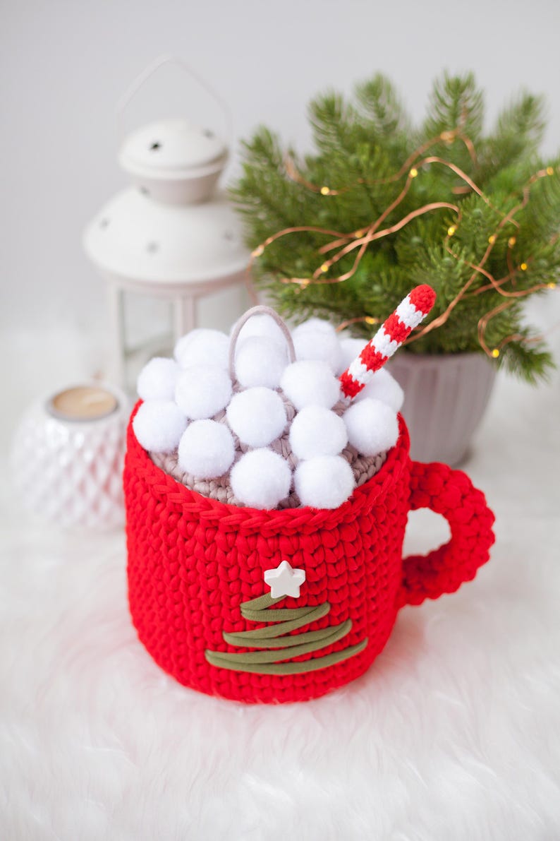 Pu&ograve; includere: Una tazza di uncinetto rossa con un top di pon pon bianco e un design di albero di Natale di uncinetto verde. Una cannuccia a righe bianche e rosse sporge dalla parte superiore.