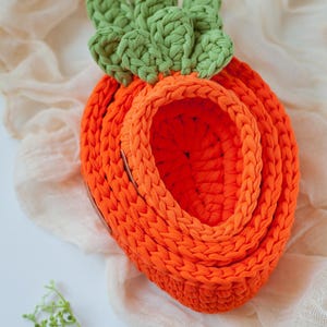 May include: A crocheted carrot-shaped basket with a green top and orange body. The basket is made of thick yarn and has a woven texture.