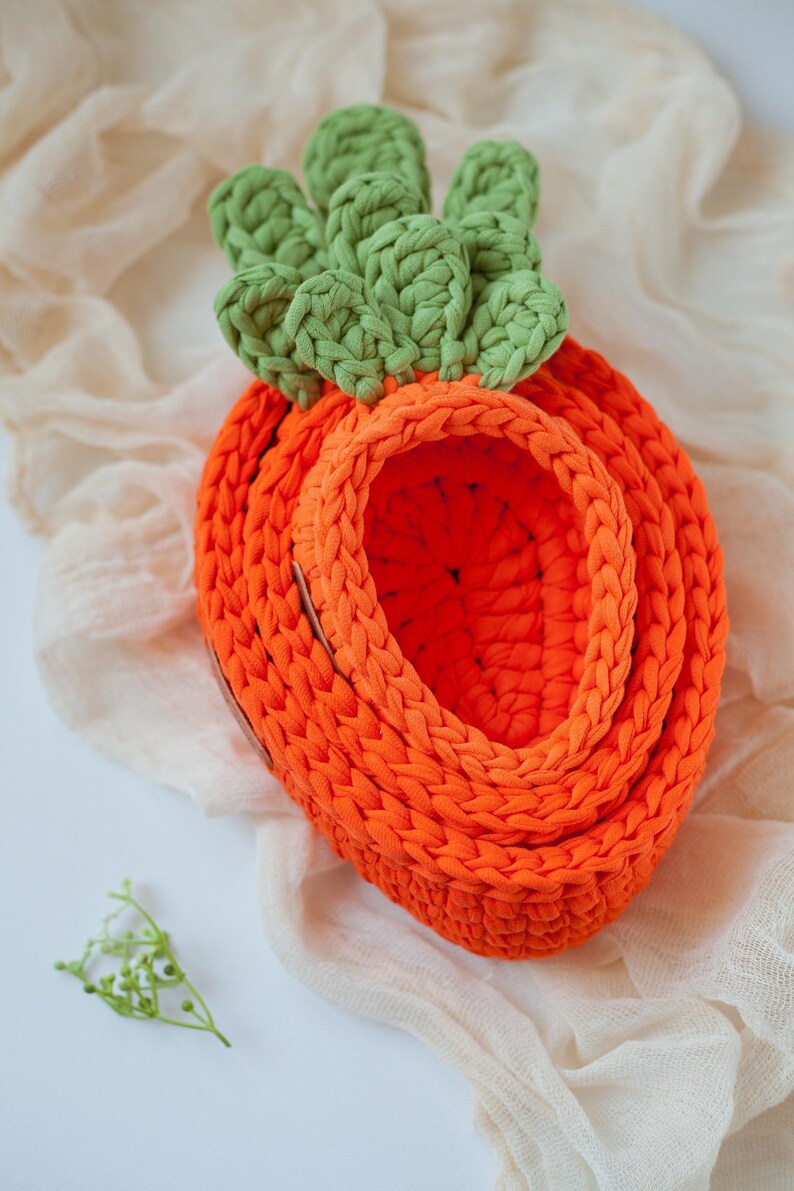 May include: A crocheted carrot-shaped basket with a green top and orange body. The basket is made of thick yarn and has a woven texture.