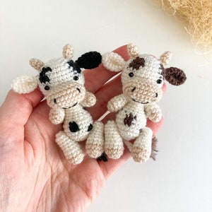 May include: Two crocheted cow plushies, one with black and white spots and the other with brown and white spots. Both have black eyes and a smiling face.