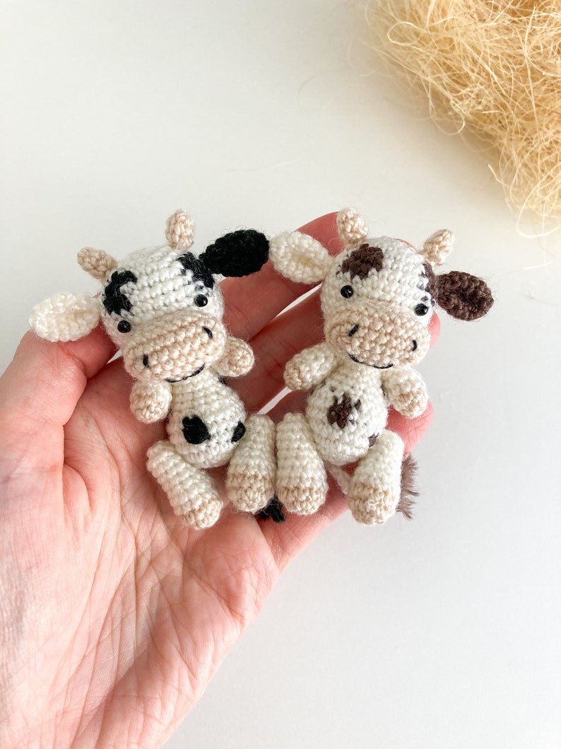 May include: Two crocheted cow plushies, one with black and white spots and the other with brown and white spots. Both have black eyes and a smiling face.