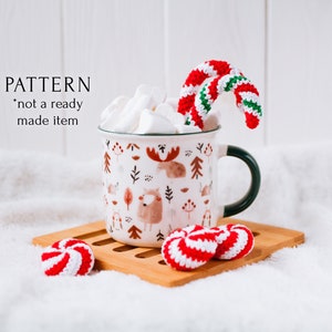 Puede incluir: Una taza de esmalte blanco con un diseño de animales del bosque, llena de malvaviscos y un bastón de caramelo de ganchillo. Dos caramelos de menta poivrée de ganchillo están sobre un posavasos de madera. El texto "PATTERN *not a ready made item" es visible.