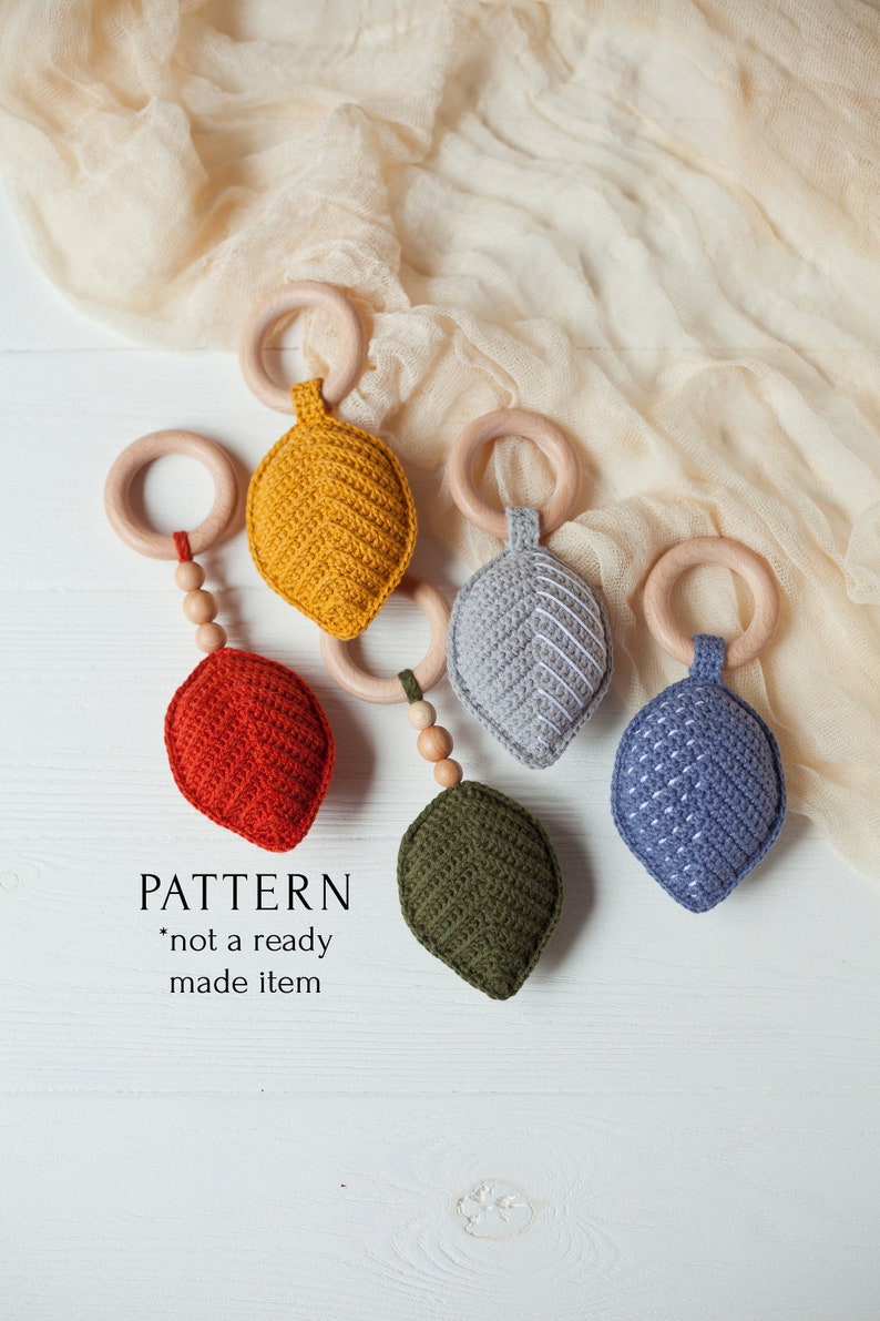 May include: Five crocheted leaf-shaped baby teethers in different colors, attached to wooden rings. The teethers are red, yellow, green, gray, and blue. The text "PATTERN *not a ready made item" is written below the teethers.