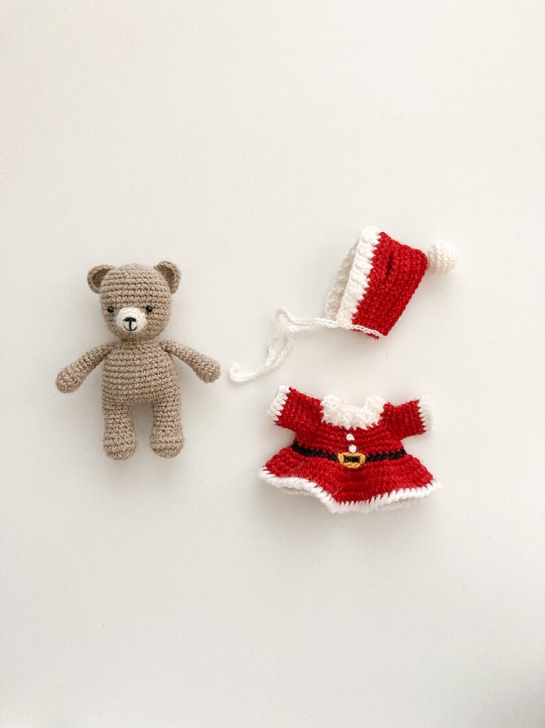 K&ouml;nnte beinhalten: Ein geh&auml;kelter Teddyb&auml;r, der eine rot-wei&szlig;e geh&auml;kelte Weihnachtsm&uuml;tze und ein rotes geh&auml;keltes Kleid mit einem schwarzen G&uuml;rtel tr&auml;gt.