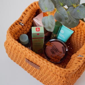 May include: A yellow crocheted basket filled with various beauty products, including a bottle of face serum, a tube of face cream, a tube of lip balm, a compact of bronzer, and a box of face cream. The basket is sitting on a white surface.