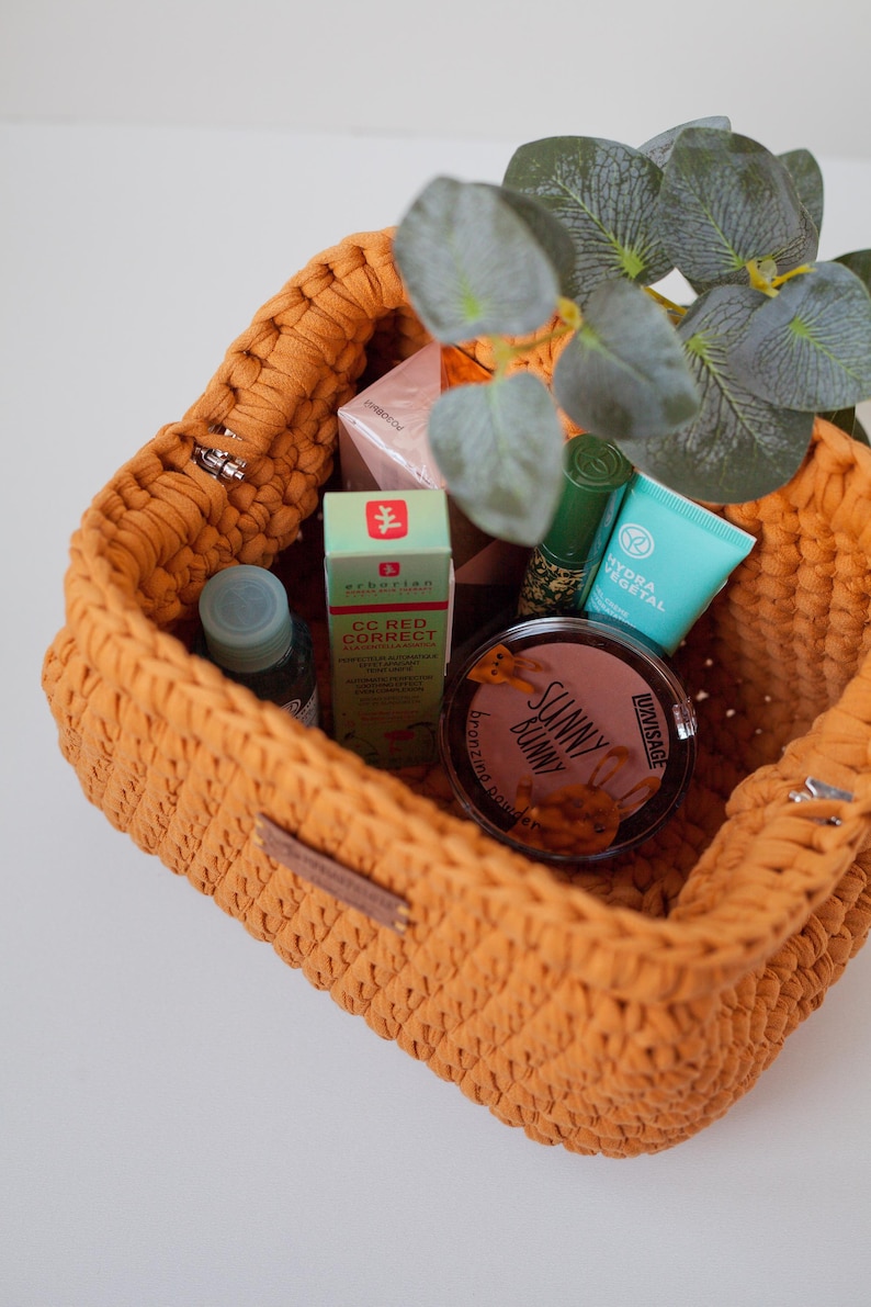 May include: A yellow crocheted basket filled with various beauty products, including a bottle of face serum, a tube of face cream, a tube of lip balm, a compact of bronzer, and a box of face cream. The basket is sitting on a white surface.