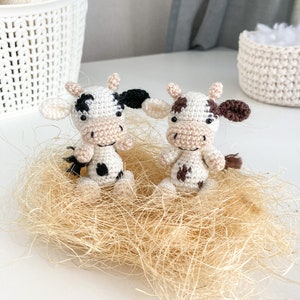 May include: Two crocheted cow plushies with black and brown spots, sitting in a nest of tan straw.